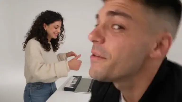 A curly-haired woman plays a small keyboard in a white studio while a man in a black jacket leans in and points