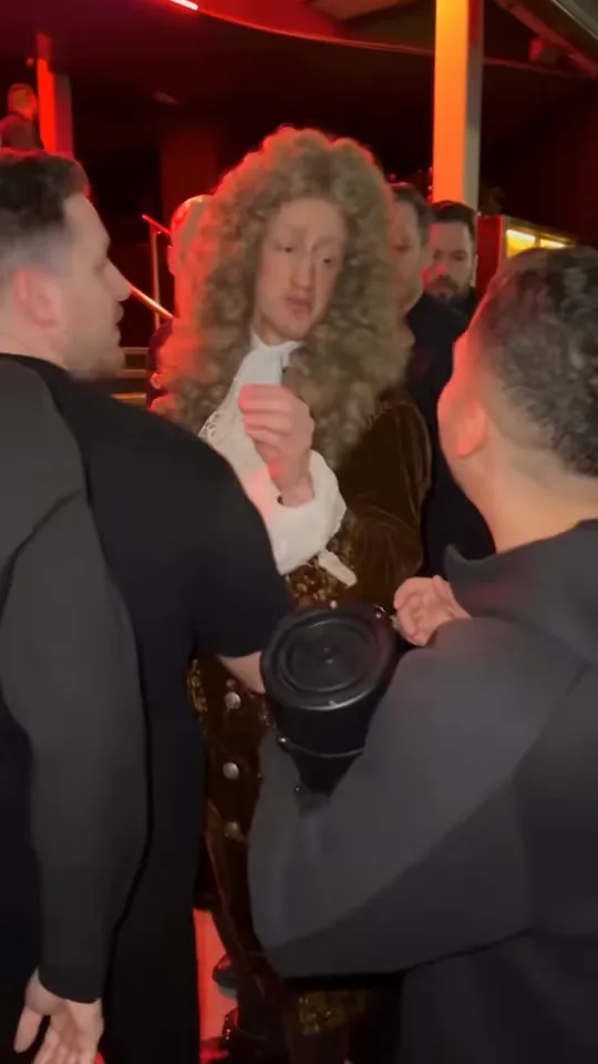 AI character in a curly baroque wig and white lace collar freestyling in a red-lit doorway at night, mouth open and one hand gesturing