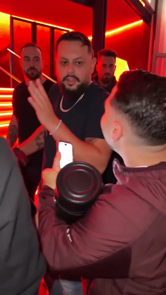 Man in a black T-shirt and gold chain pointing at the camera in a red-lit doorway, a portable speaker held up in the foreground