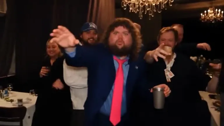 A curly-haired man in a navy suit and pink tie leads a cheering crowd under crystal chandeliers, one arm reaching out and a silver cup in the other hand