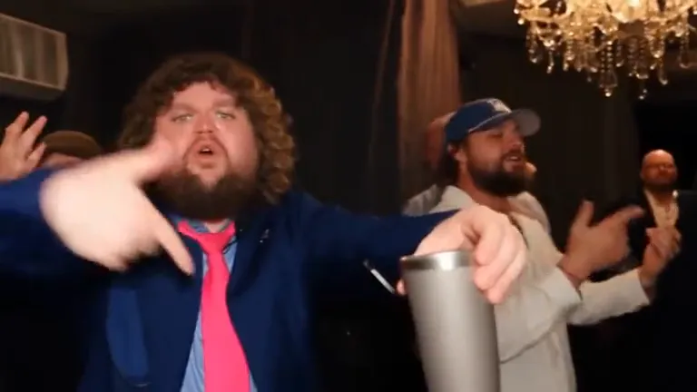 Close-up of the same man holding a silver cup up and pointing at the camera, a chandelier and a friend in a cap behind him