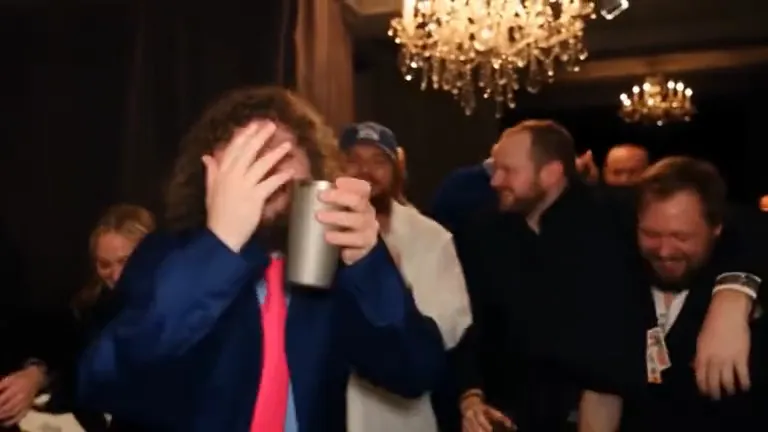 The same man puts one hand flat over his eyes and searches the room while holding a silver cup, friends doubled over laughing behind him