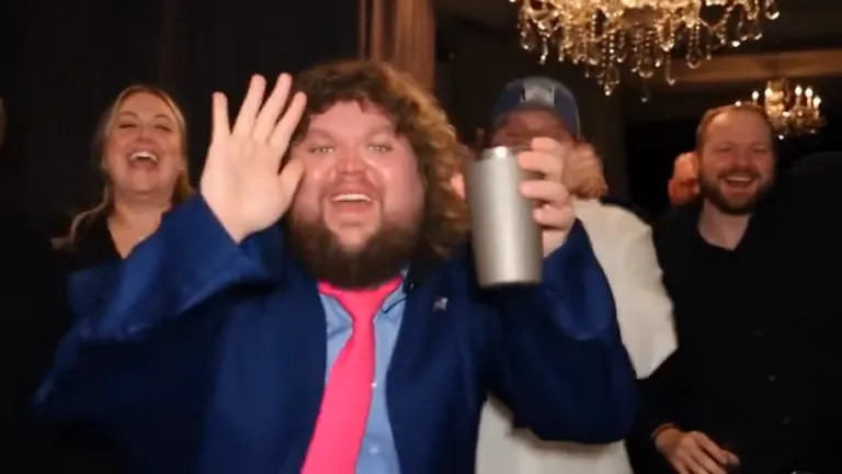 The same man drops his hand and laughs into the camera, waving and raising a silver cup as the crowd behind him cheers