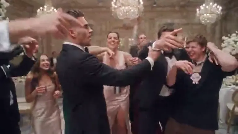 A young man in a black tuxedo shakes a silver cup in front of a cheering crowd in a ballroom of crystal chandeliers and white flowers