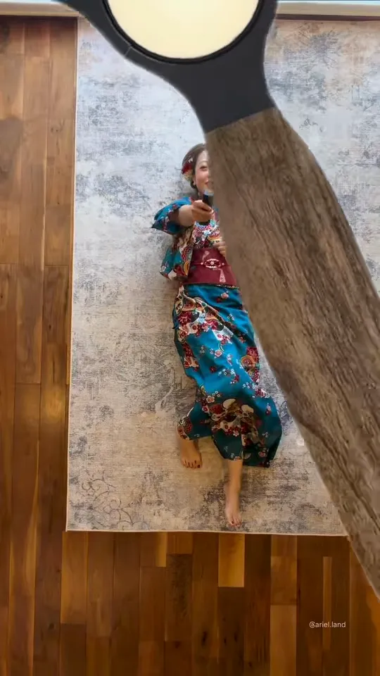 Overhead view of a woman in a blue floral kimono lying on a grey rug as a wooden ceiling fan blade sweeps across from the right