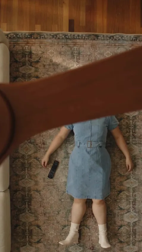Overhead view of a woman in a blue denim shirt-dress and white socks on a patterned rug as a brown wooden fan blade sweeps over her