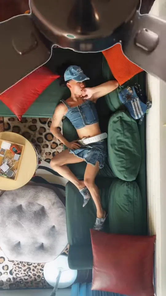 Overhead view of a man in a denim vest, denim shorts and cap lying sideways on a green sofa with a dark fan blade at the top