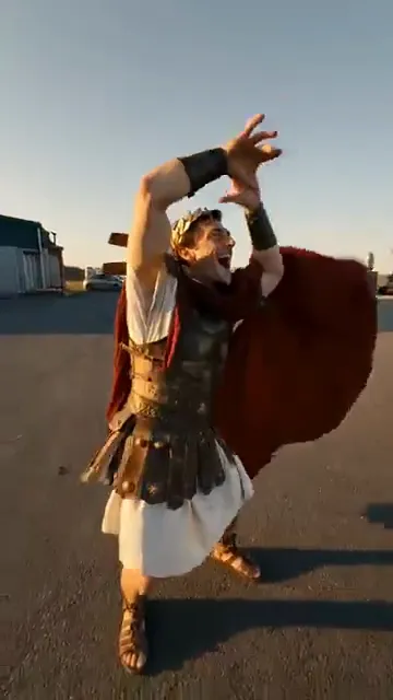 An AI-made Roman emperor in a red cape throws both arms up high on an empty lot at sunset