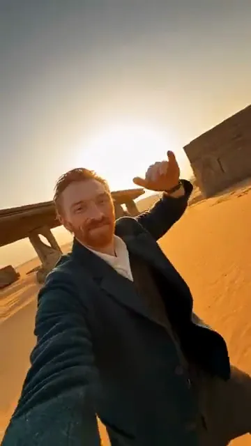 An AI-made painter in a dark coat holds the phone at arm's length and points back over the shoulder in a desert selfie