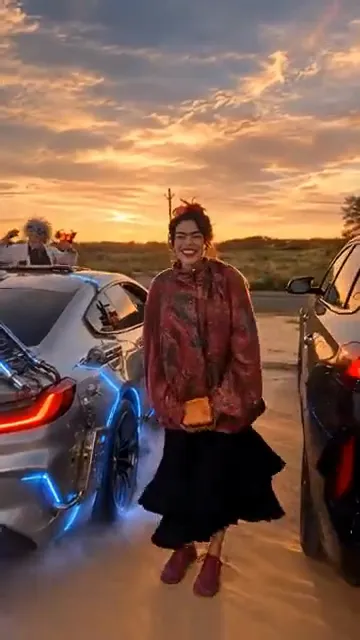An AI-made painter in a red shawl smiles beside a sports car turned into a glowing blue time machine under a sunset sky