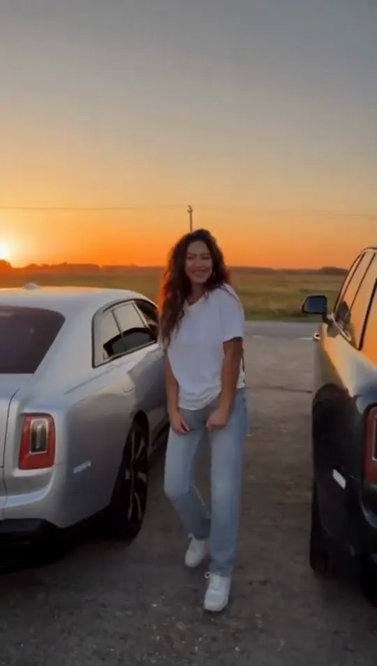 A woman in a black tracksuit throws both arms up and laughs on an empty gas station lot at dusk, trucks and a red car behind her