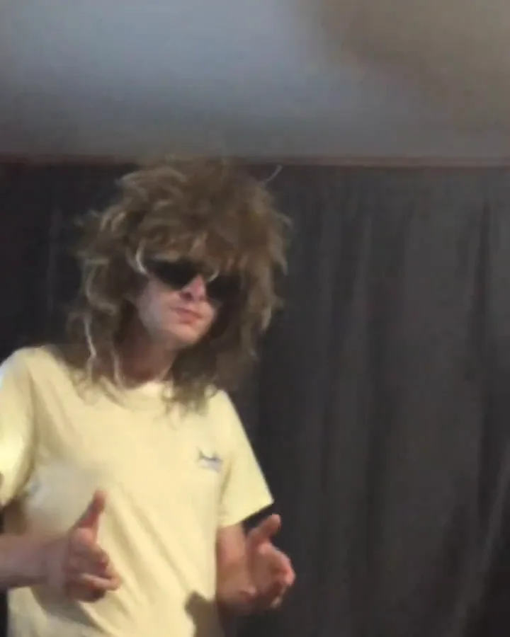 A long-haired person in a wig and dark sunglasses makes finger guns in front of a dark curtain