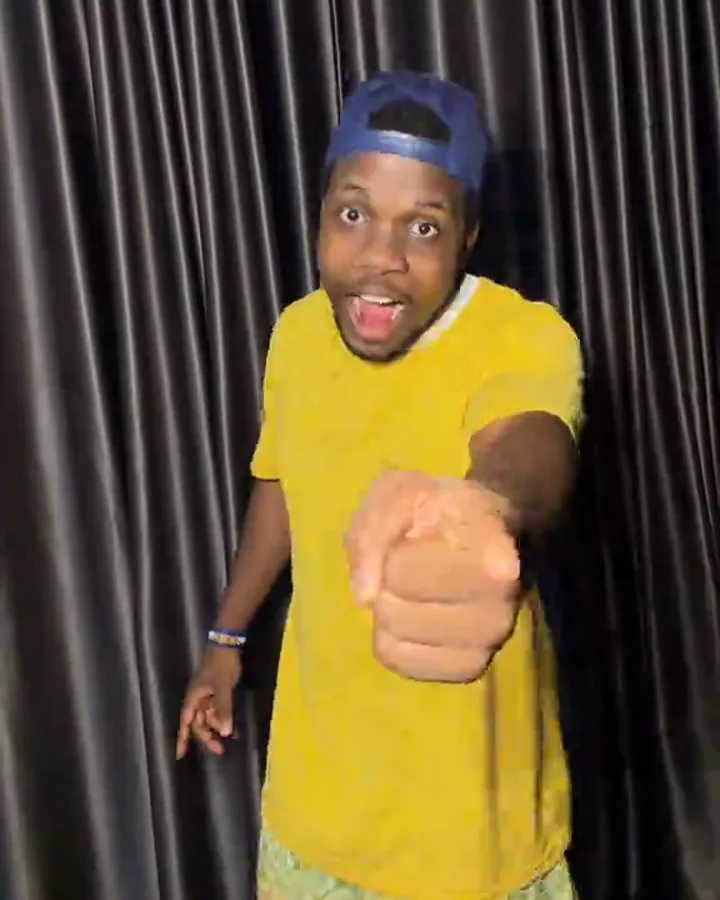 A man in a yellow T-shirt and a blue cap points a finger at the lens in front of a dark satin curtain