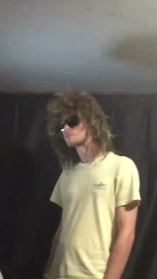 A person in a long wig, dark sunglasses and a pale yellow T-shirt lifts the chin in front of a dark curtain