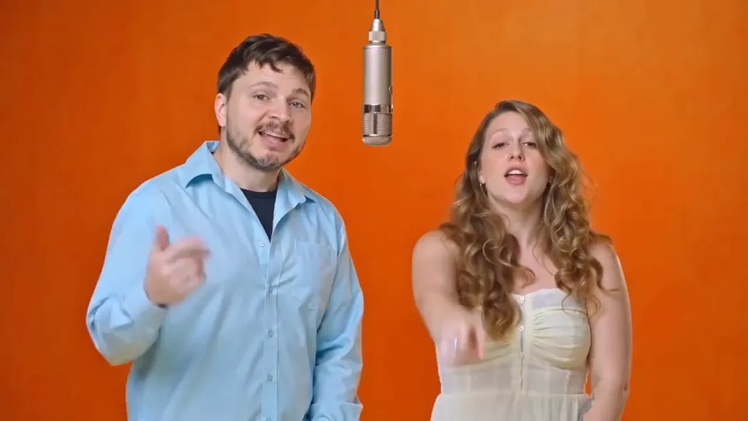 Opening beat of a hotel lobby AI video: two people in front of a plain orange wall with a single hanging microphone between them, he gestures while she points at the camera