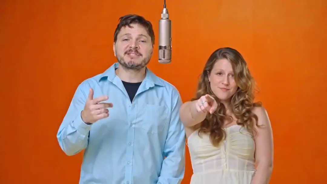 Both people point straight at the camera in sync under the hanging microphone against the orange studio wall