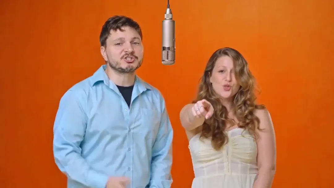 The person on the left raps facing the camera while the person on the right points at the lens, orange wall and hanging mic behind them