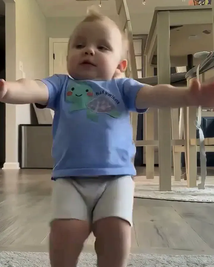A baby in a blue tee and shorts flinging both arms out wide like wings