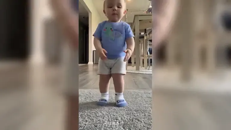 A baby in a blue tee and grey shorts standing on a grey rug, ready to dance