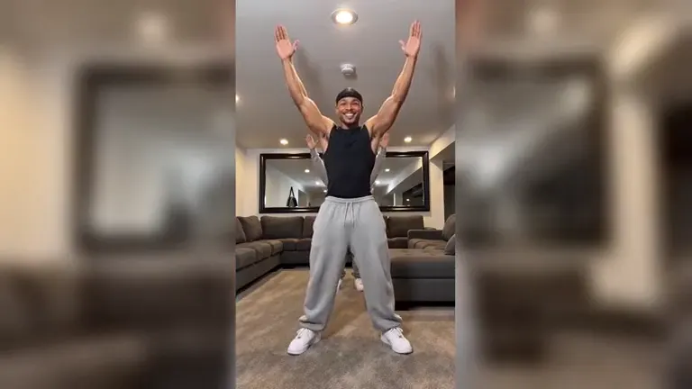 A man in a durag, black tank top and grey sweatpants in a basement with a grey sectional sofa, arms up in a V