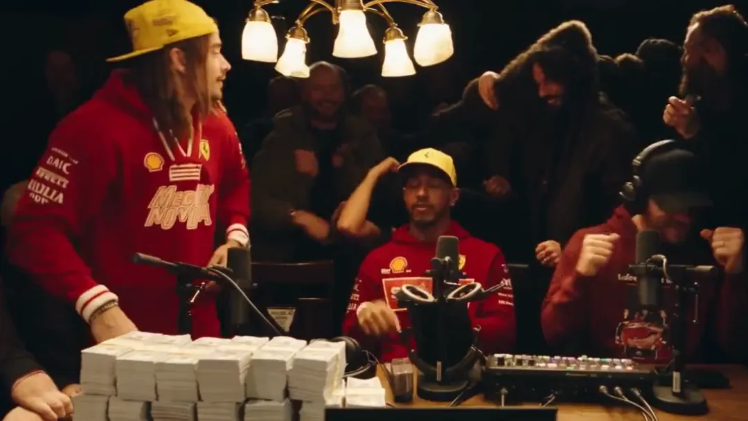 Two performers in red jackets and yellow caps at a long wooden table piled with stacks of cash, one standing and one seated, a crowd behind them under a chandelier