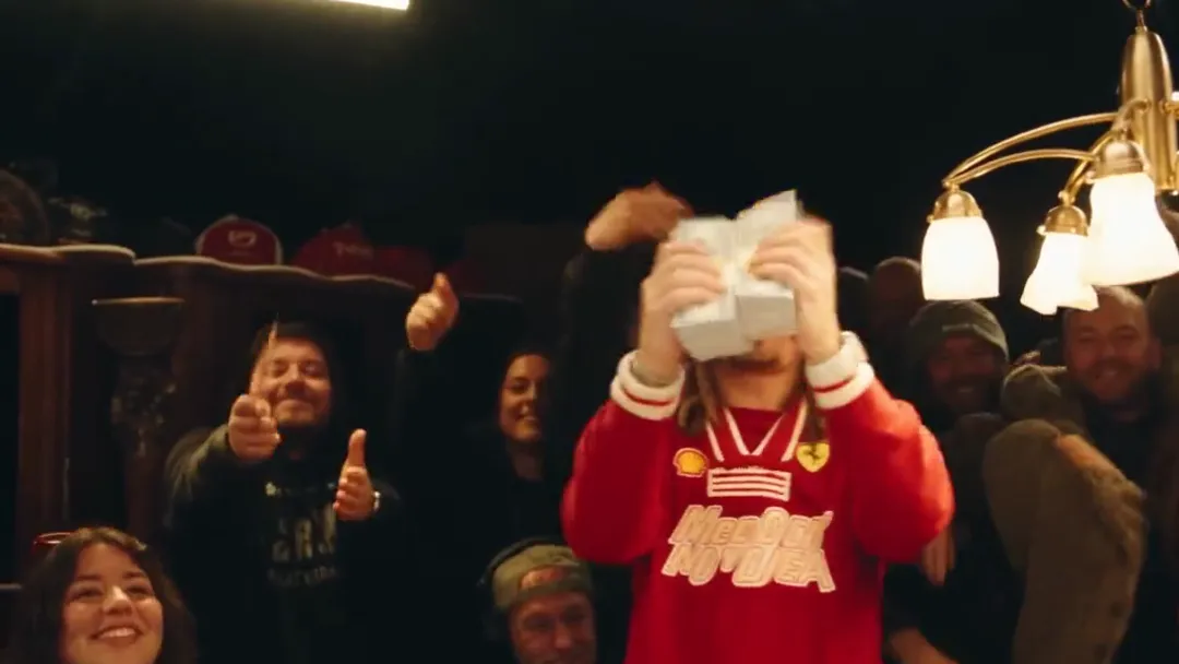 A performer in a red jacket holds two stacks of cash up in front of his face while people in the crowd behind him grin and give thumbs up