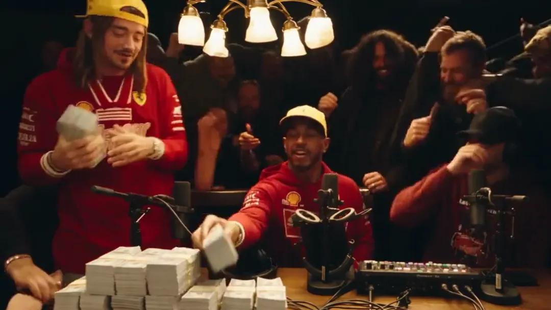 The standing performer holds one stack of cash while the seated performer slaps another stack down on the table and the crowd raises hands