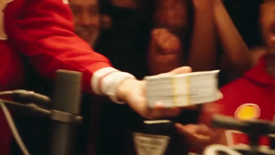 Close-up of a hand in a red sleeve holding a banded stack of cash out across the table, faces out of focus behind it