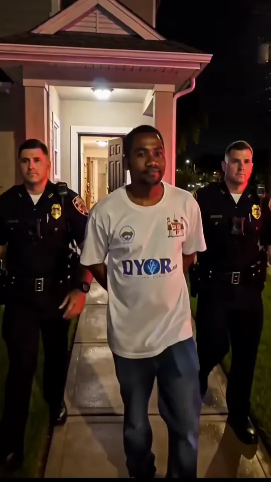 A man in a white T-shirt and jeans is walked down the front path of a house at night by two officers, porch light on behind them