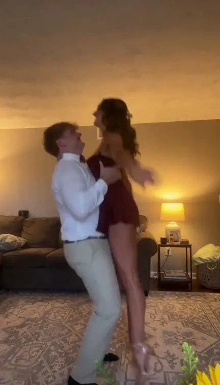 A young man in a white shirt and khaki trousers lifts a young woman in a short burgundy dress off a patterned rug in a lamp-lit living room (homecoming outfits).