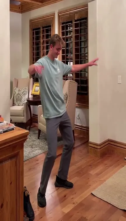 A young man in a teal t-shirt and grey trousers with arms out on a wooden floor in a living room.