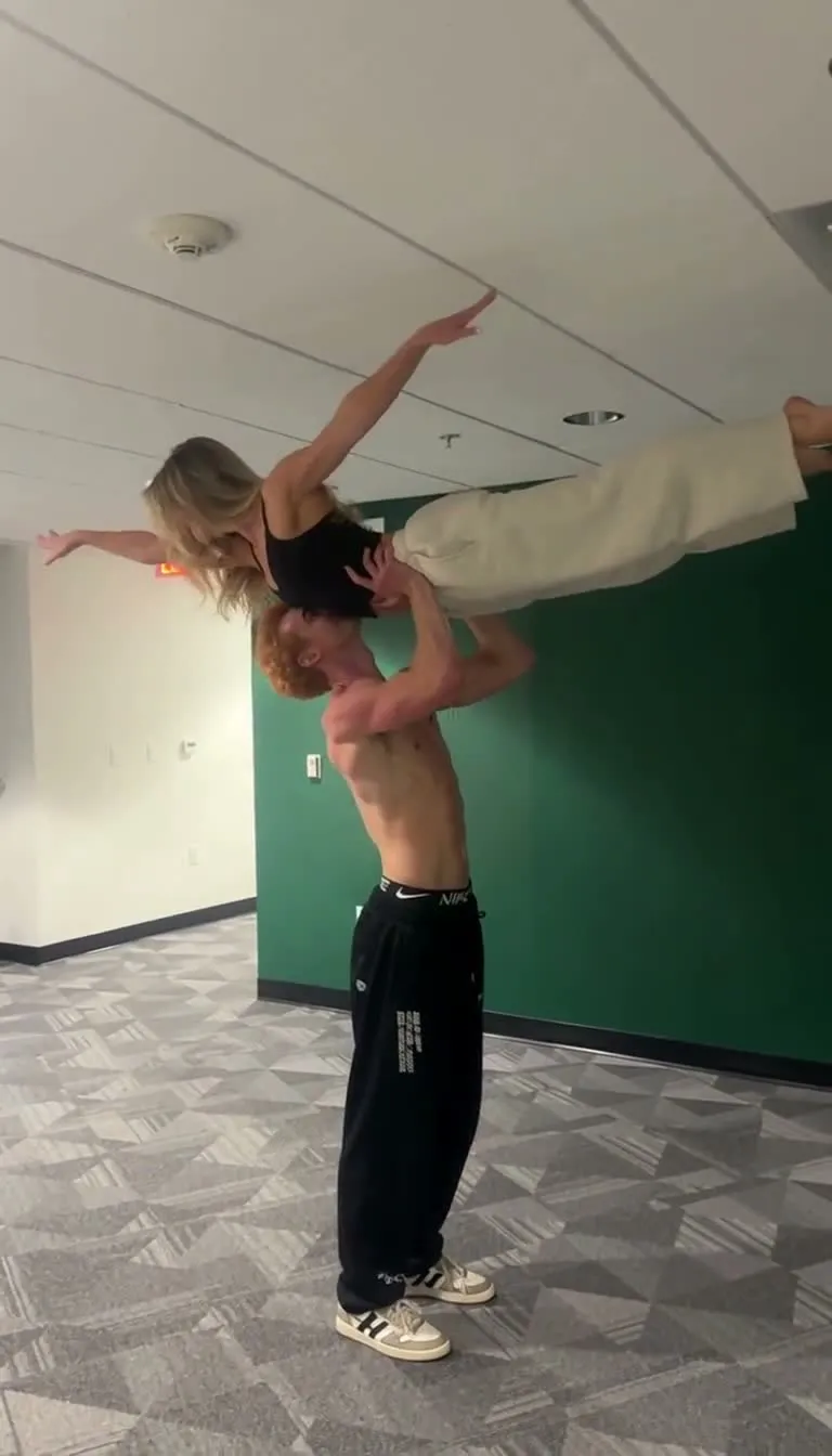 The same room, he holds her flat overhead with her arms out.
