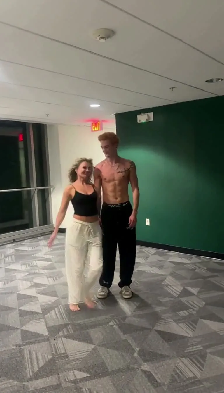 A young woman in a black top and cream trousers stands beside a shirtless young man in black trousers in a bright empty room with a green wall, before the lift.