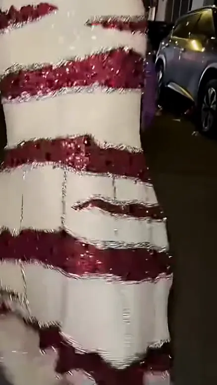 Shot one of a ramalama costume walk: a low, shaky close-up of a white-and-burgundy sequined gown walking down a night street, a purple glove at the edge of the frame, no face yet