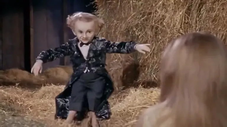 A small figure in a black velvet tailcoat and white bow tie tiptoes across the hay in a dim barn, knees bent and arms flung wide, while a blonde woman watches from the foreground