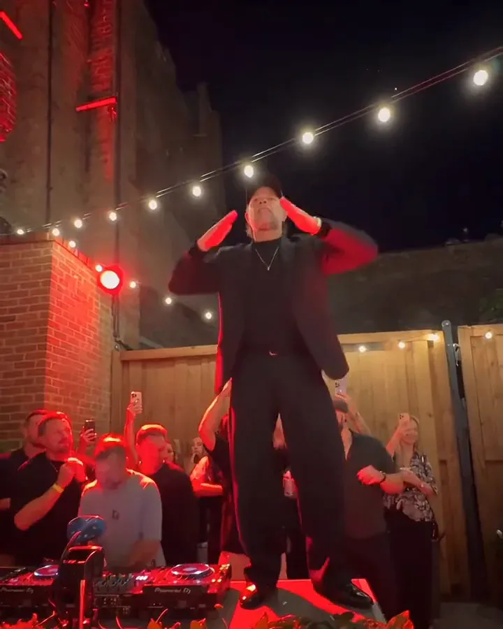 A man in a black suit snaps both hands up to the sides of his head
