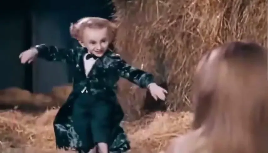 The little man opens his arms in the barn while a blonde woman watches in the foreground