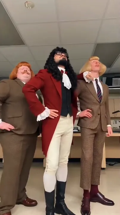 Three costumed dancers in bob wigs and retro suits stand in a row in an office kitchen with hands on hips