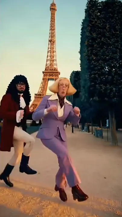 A dancer in a blond bob wig and lilac suit steps forward on a gravel path near the Eiffel Tower with a partner in a red coat behind