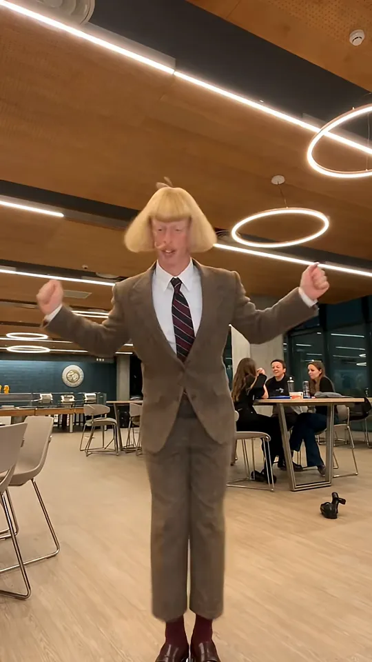 A dancer in a golden-blond bob wig and brown tweed suit raises both fists in a cafeteria with ring lights while people sit at a table behind him