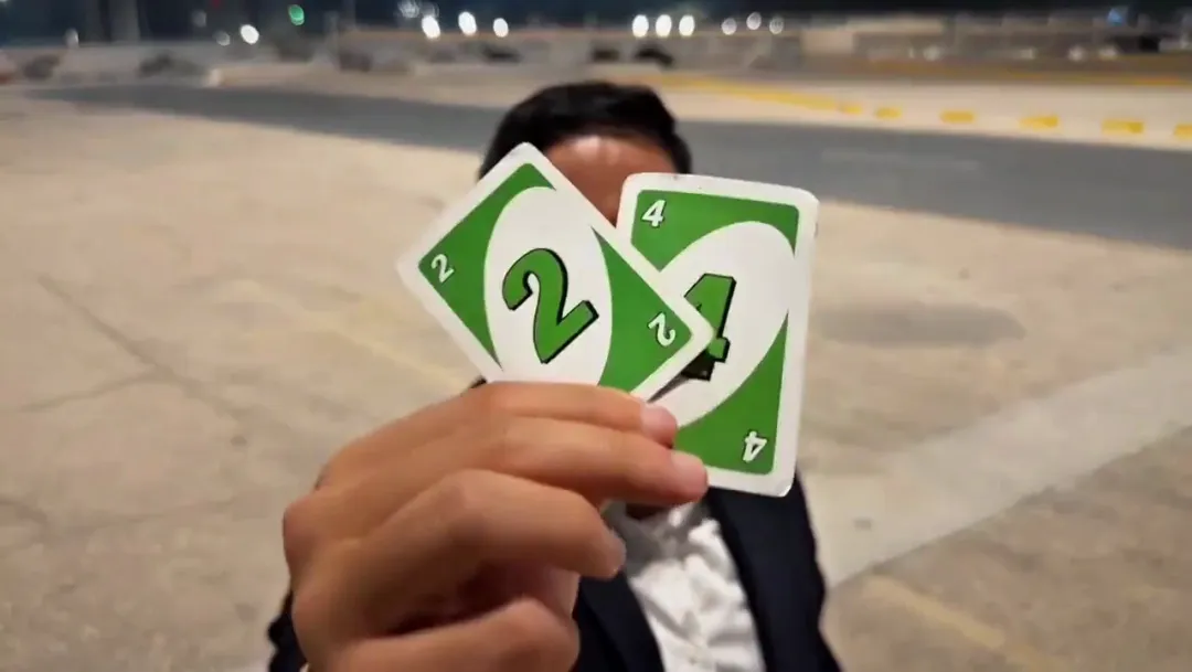 A man in a dark suit holds a green 2 card and a green 4 card over his face on an empty lot at night