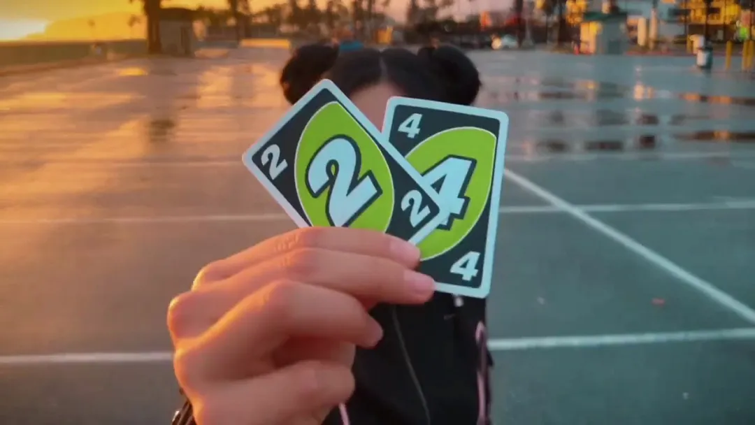 A woman in silver wraparound sunglasses holds a green 2 card and a green 4 card up to the lens in a car park at sunset