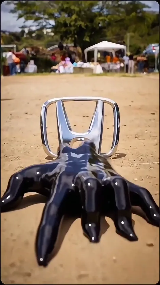 A black liquid hand closes over a car badge lying on a sunny dirt lot at a car meet