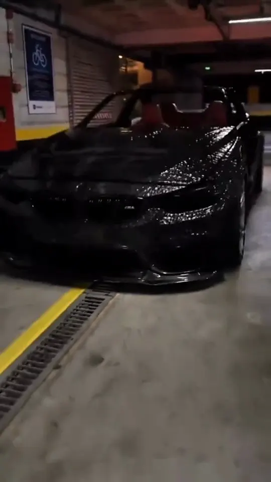 A black convertible coupe, just rebuilt from black liquid, sits in an underground car park beside yellow lane lines and a drain grate