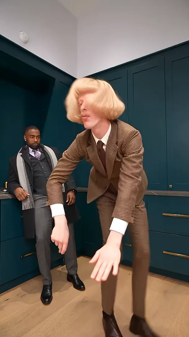 In a kitchen with dark green cabinets, a man in a brown suit bends deep forward and swings both hands at the floor while a friend leans on the counter and watches