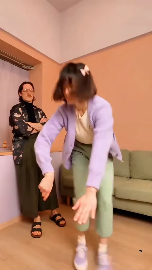 In a pink living room, a woman in a lilac cardigan leans forward and swings her arms low while a friend stands behind with arms crossed