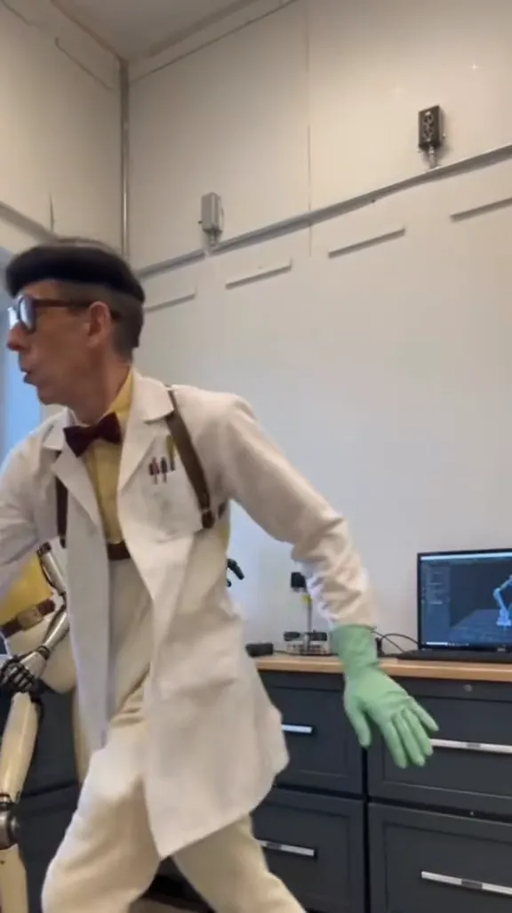 In a lab with grey cabinets and monitors, a man in a white lab coat and green gloves dances low next to a robot